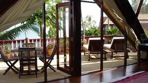 A room with a wooden floor, a bed with a white and red bedspread, a window with a view of the sea and palm trees, and a balcony with outdoor furniture. - luxury in paradise (Koh Samui)