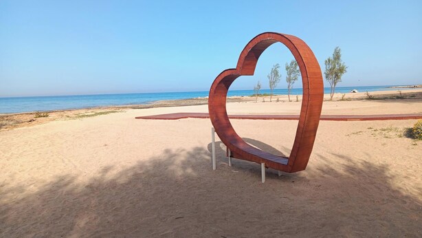 Escultura de metal em formato de coração numa praia arenosa, com um céu azul límpido e mar calmo ao fundo.