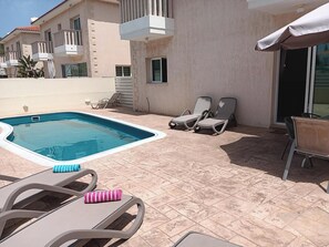 A swimming pool with lounge chairs, a covered patio, and a building with balconies.