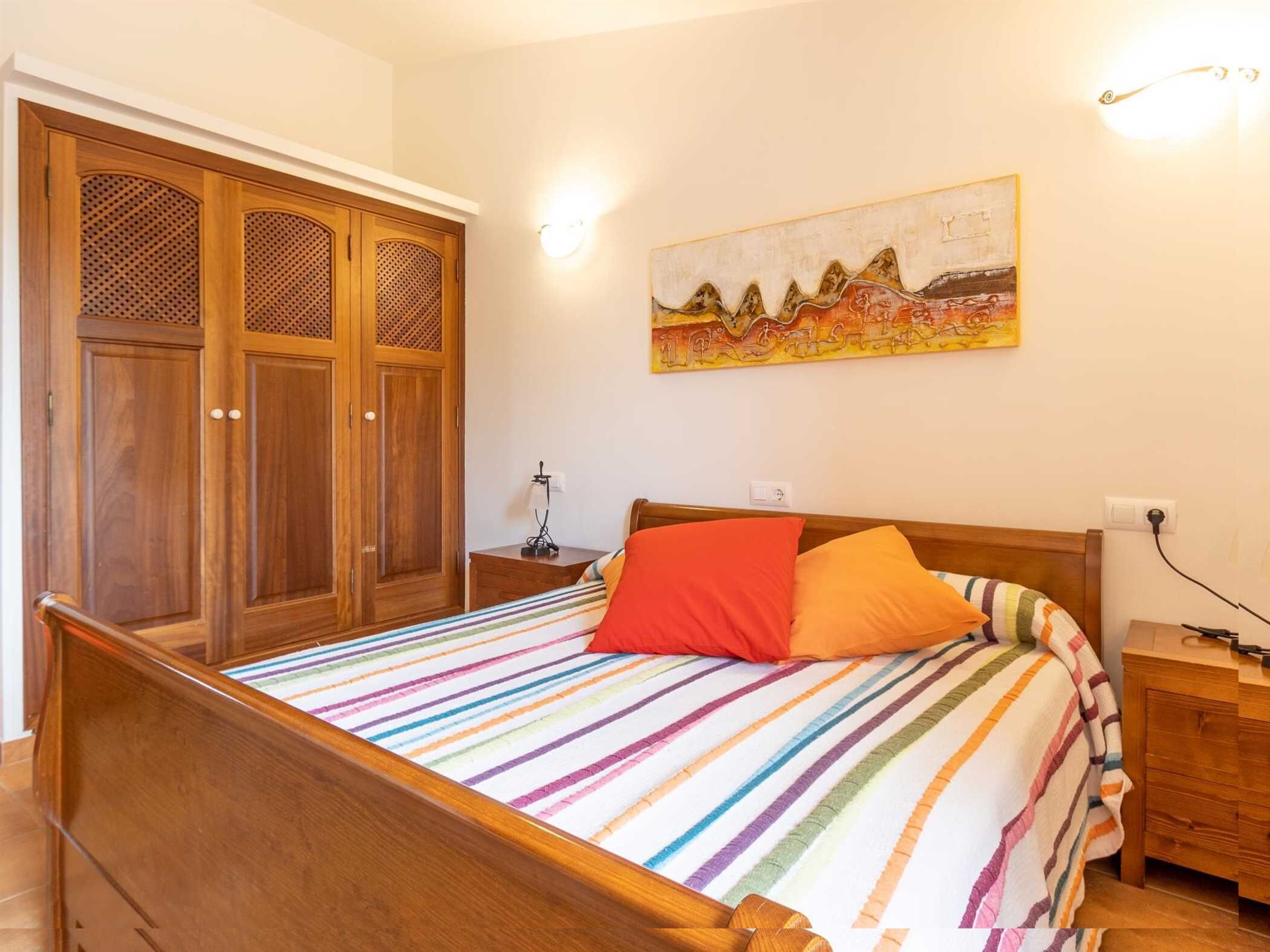 Een slaapkamer met een houten bed, kleurrijke strepenbeddengoed, een nachtkastje, een lamp en een kledingkast met decoratief paneel.