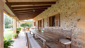 A covered outdoor dining area with wooden benches and a long table, stone walls, and windows with shutters.