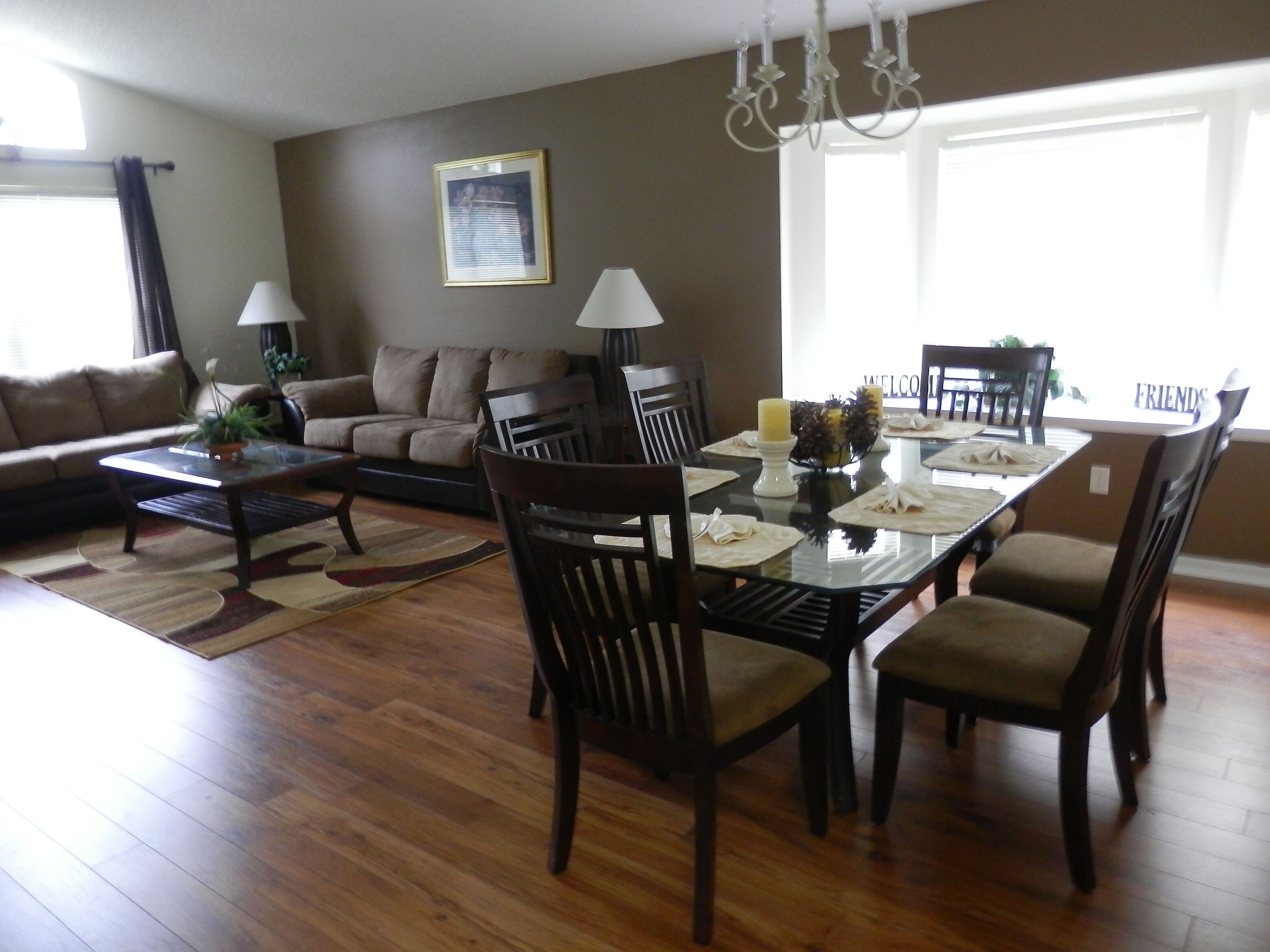 A dining area with a glass table, wooden chairs, and a chandelier. There is a sofa and a lamp in the background.