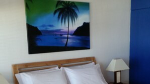 A neatly made bed with white linens, two bedside lamps, and a painting of a palm tree on the wall.