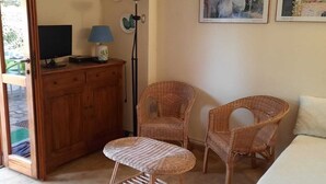 Een kamer met een bed, twee rieten stoelen, een tafeltje, een bureau met computer en lamp, en een deur naar buiten.