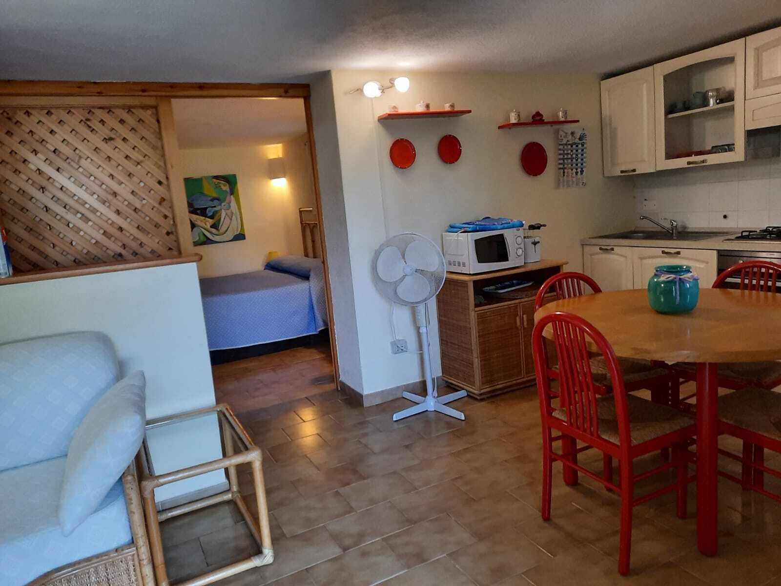 A compact living space with a bed, a dining table with chairs, a kitchenette, and a fan.