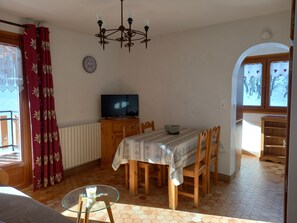 A dining area with a table, chairs, a chandelier, a television, and a view of the outside through a window. - At Riton app. 3 pers. 3 *** quiet, close to shops, at the foot of the slopes (Valloire)