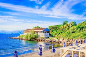 En fastighet vid kusten med ett stort hus, en strand med parasoller och utsikt över havet och bergen.