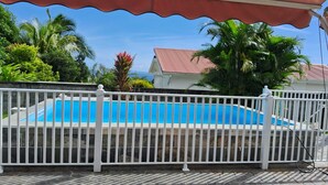 Una piscina con cerca blanca, rodeada de abundante vegetación y una casa de techo rojo.