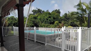 Un área de piscina con valla blanca, suelo de piedra y abundante vegetación al fondo.
