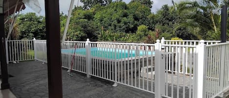 A pool area with a white fence, stone flooring, and lush greenery in the background.