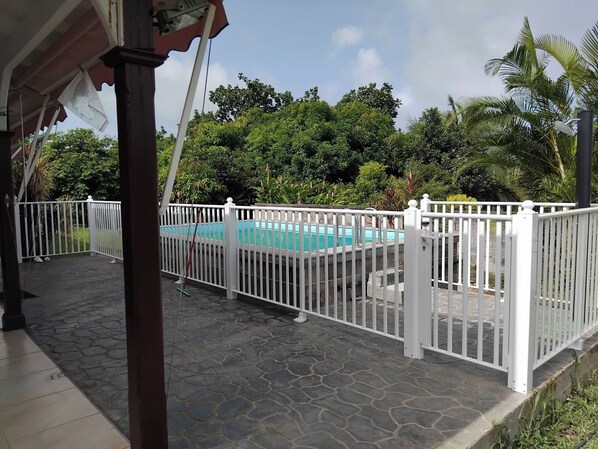 A pool area with a white fence, stone flooring, and lush greenery in the background.