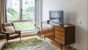 A living room with a brown leather sofa, a wooden cabinet with a television, a green rug, and a window with a view of greenery and buildings. - CdC II Apartments By Tale House (Porto)