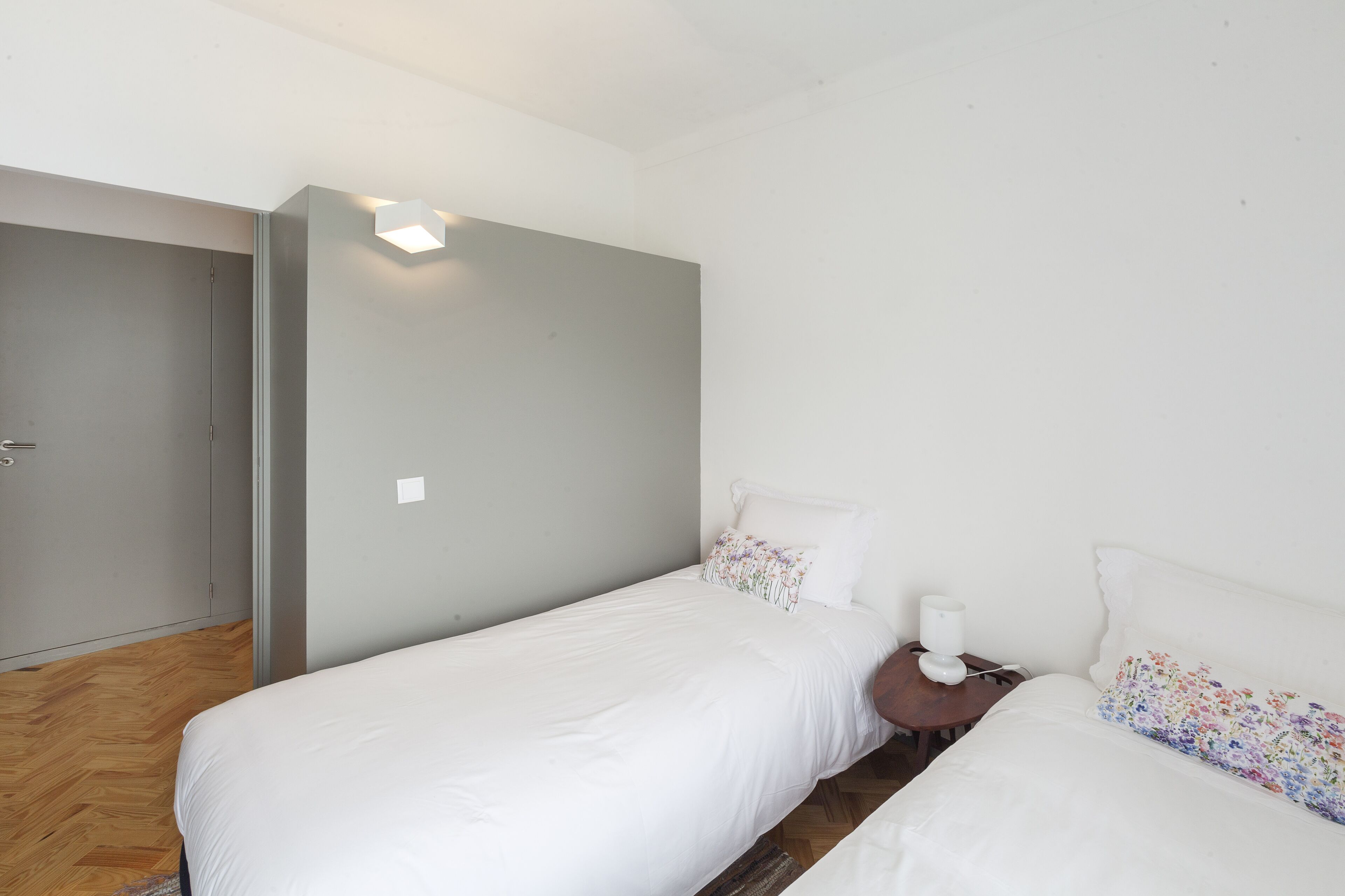 A bedroom with two single beds, a wooden floor, a grey wardrobe, and a white wall with a light fixture.