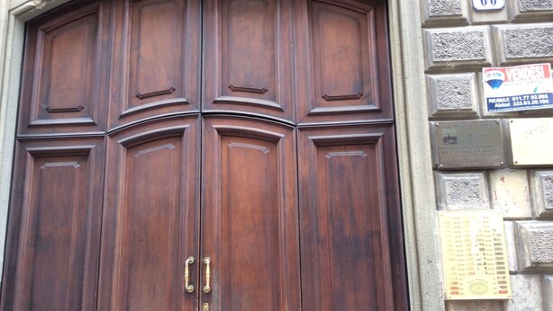Uma grande porta dupla de madeira com puxadores de latão, embutida numa parede de pedra com uma placa de número e um letreiro comercial.