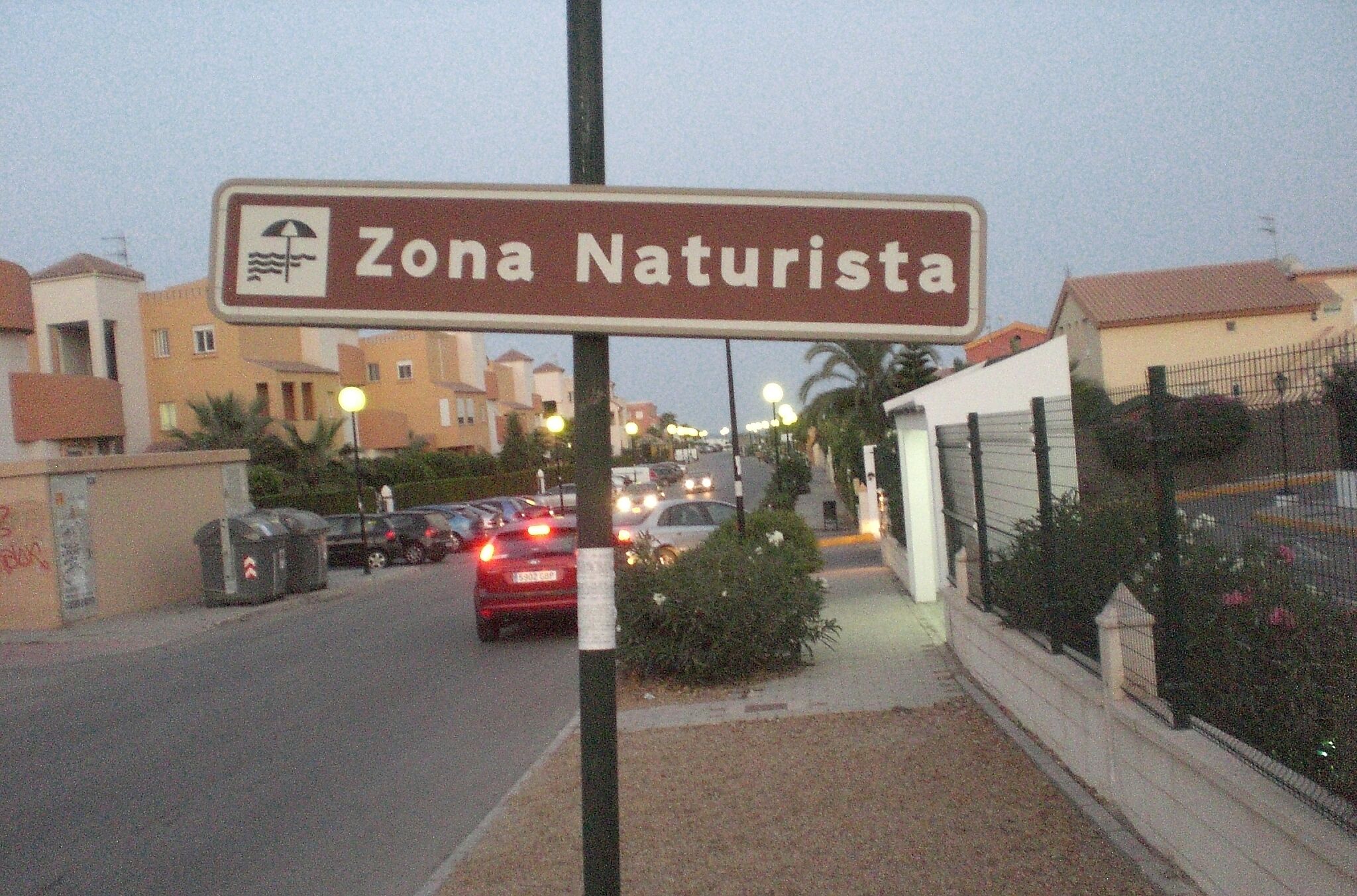 Un panneau de signalisation indiquant « Zone naturiste », avec un symbole représentant un parasol de plage, entouré de bâtiments et de voitures garées.