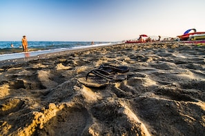 Un par de chanclas sobre una playa arenosa, con gente y sombrillas de playa al fondo.
