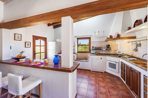 Une cuisine avec un plafond orné de poutres en bois, des murs blancs et un mur recouvert de carreaux. On y trouve des meubles blancs, un évier et une fenêtre donnant sur un jardin.
