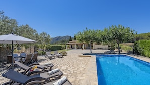 Ein Poolbereich mit Liegestühlen und einem Tisch unter einem Sonnenschirm, umgeben von Grünflächen und einer Steinterrasse.