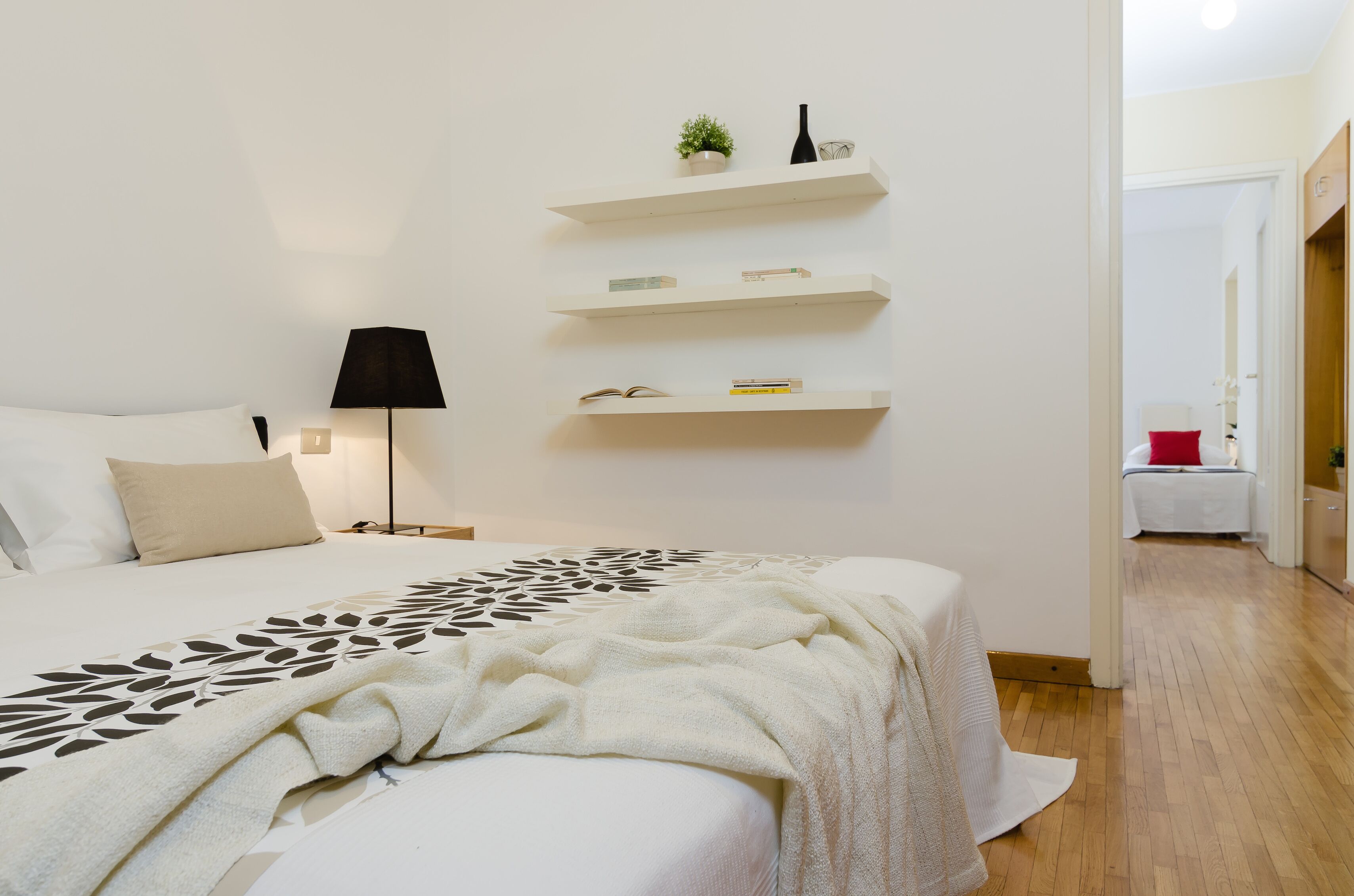 A bedroom with a bed, bedside lamp, and wall-mounted shelves with books and a plant.