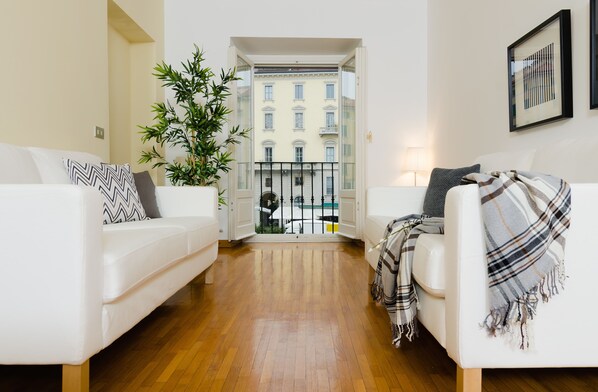 A modern living room with two white sofas, a large potted plant, and a view of a building through an open door.
