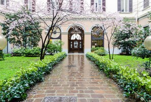 A stone pathway leading to a building with a central door, flanked by green hedges and flowering trees.