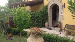 A yellow building with a stone archway entrance, a large vase with flowers in the foreground, and a bench to the right. - Country Cottage / Gite - Pergola (Pergola)