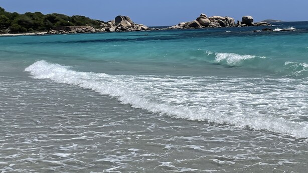 Une plage avec une eau turquoise et limpide, de douces vagues et des formations rocheuses en arrière-plan.