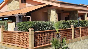 A two-story house with a stone facade, a tiled roof, and a fenced courtyard with a palm tree. - WELL-LIT MEDITERRANEAN VILLA WITH VERANDA AND GARDEN ONLY 50 METERS FROM THE SEA (Campofelice PA, Italia)