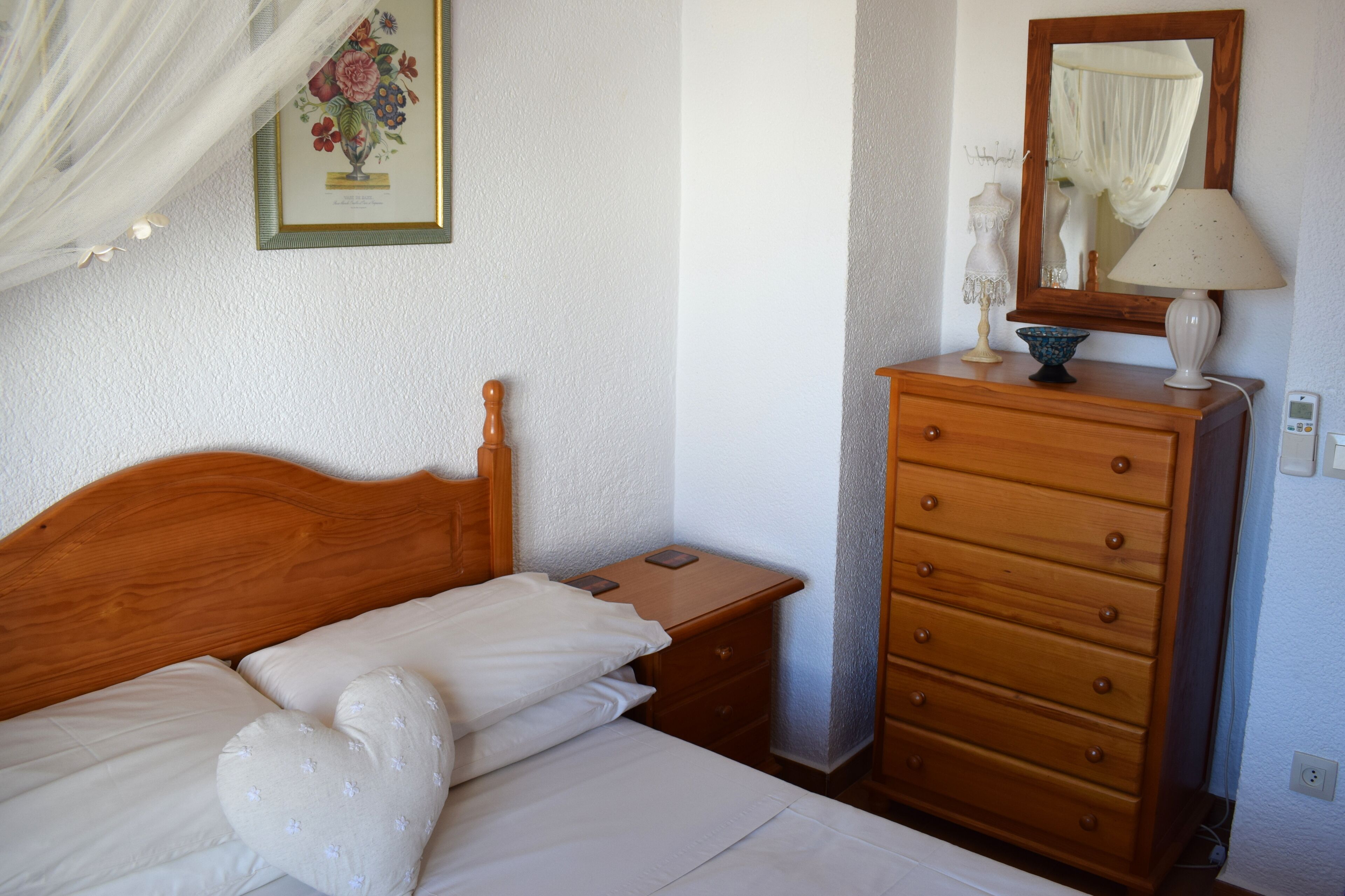 A neatly made bed with white linens and heart-shaped pillows, a wooden nightstand with a lamp, a dresser with drawers, and a framed floral artwork on the wall.
