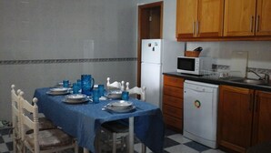 Un coin repas avec une nappe bleue, des chaises blanches, un réfrigérateur, un four à micro-ondes et des meubles de rangement en bois.
