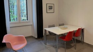 Un coin repas comprenant une table, des chaises, un fauteuil à bascule, une fenêtre avec des rideaux et un tableau accroché au mur.