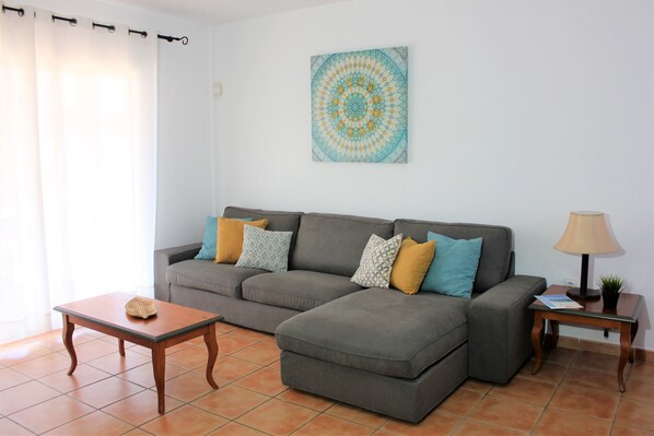 A living room with a grey sectional sofa, a small wooden table, a lamp, and a wall-mounted tapestry.