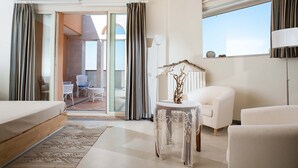 Quarto de hotel com cama, área de estar com cadeiras, mesa com objeto decorativo e uma grande janela com cortinas.