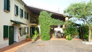 A courtyard with a cobblestone path, a covered outdoor seating area, and a building with green shutters and a balcony. - Casale Gelsomino First floor (ROVOLON)