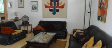 Una sala de estar con dos sofás negros, una mesa de centro de vidrio y varios cojines. Hay cuadros enmarcados en las paredes y un cuadro abstracto grande sobre la mesa de centro.