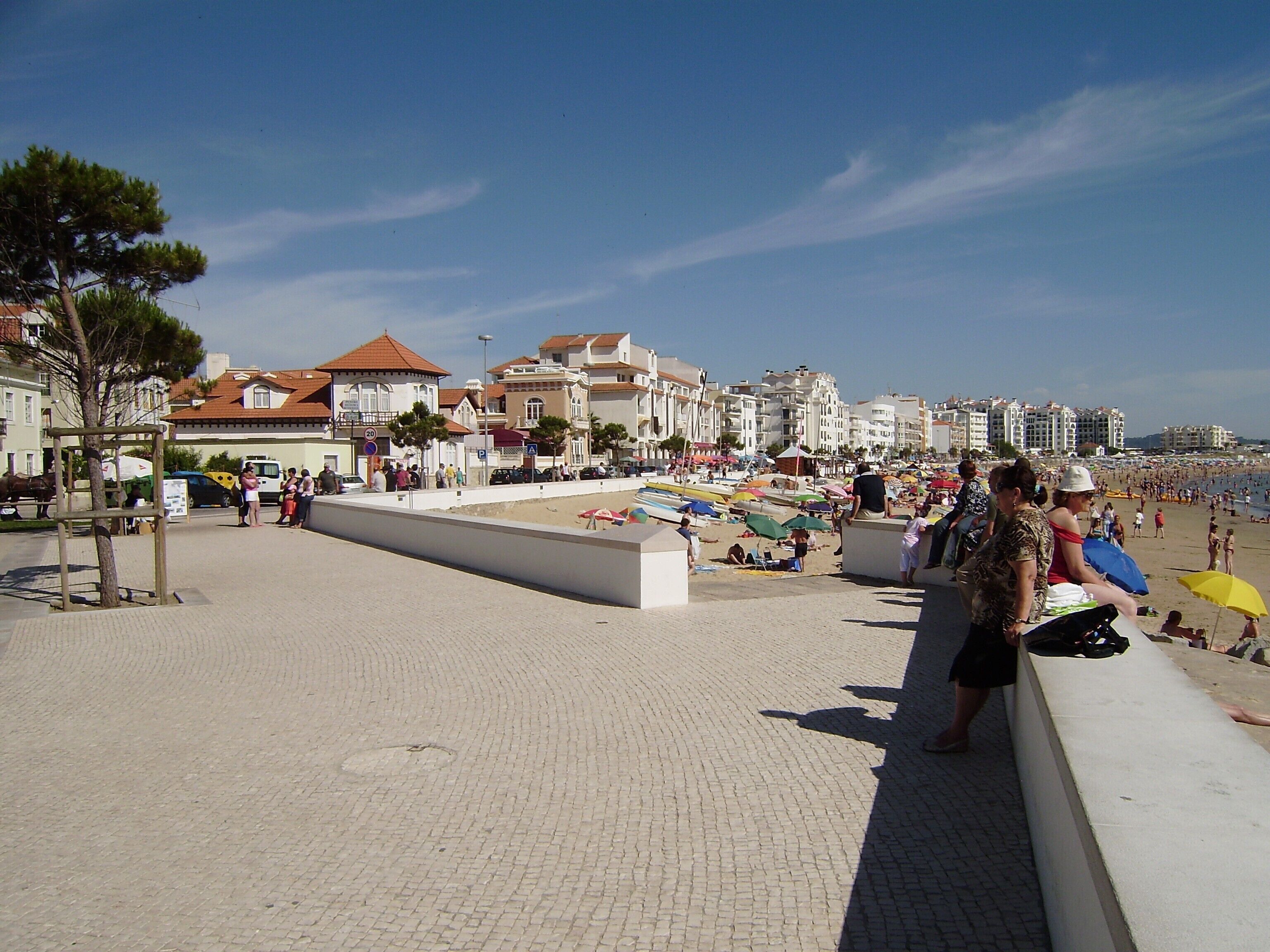 A paved promenade with buildings in the background, a clear sky, and people on the beach.