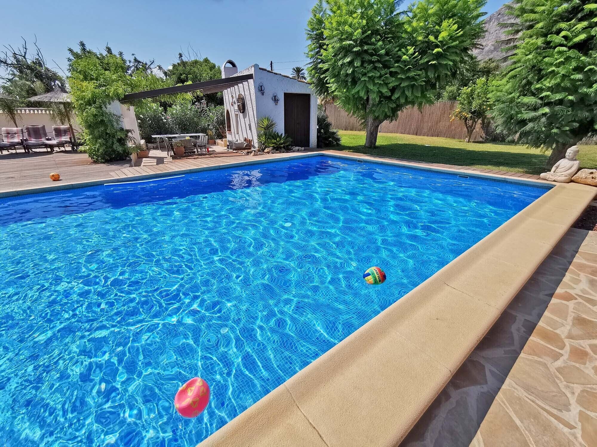 A clear blue swimming pool with a stone border, surrounded by a patio area with outdoor furniture and a small building.