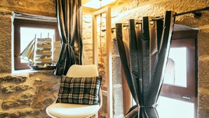 A room with stone walls, a wooden floor, a white chair with a plaid pillow, and a window with curtains. - Charming Gite in Medieval Village 1mn walk to Shops & Restaurants (Nolay)