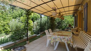 An outdoor patio with a table and chairs, surrounded by greenery and a covered area with hanging plants. -  part of villa on the ground floor next to a vineyard (Lorgues)