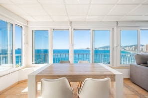 A spacious room with a large wooden table and white chairs, large windows with a view of the sea and cityscape.