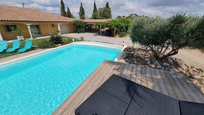 A rectangular swimming pool with a wooden deck and a pergola with a table and chairs.