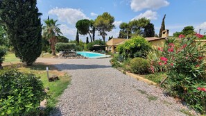 A gravel pathway leading to a swimming pool surrounded by lush greenery and a building with a tiled roof.