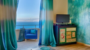 A room with a view of the sea, a blue sofa, a television on a cabinet, and a patterned cabinet.