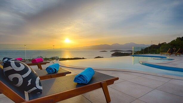 A poolside area with lounge chairs and towels, overlooking a body of water and distant mountains during sunset.
