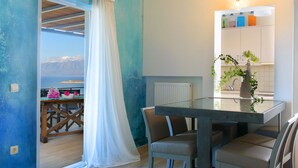 A dining area with a table and chairs, a view of the sea through a window, and a kitchen area with cabinets and a plant.