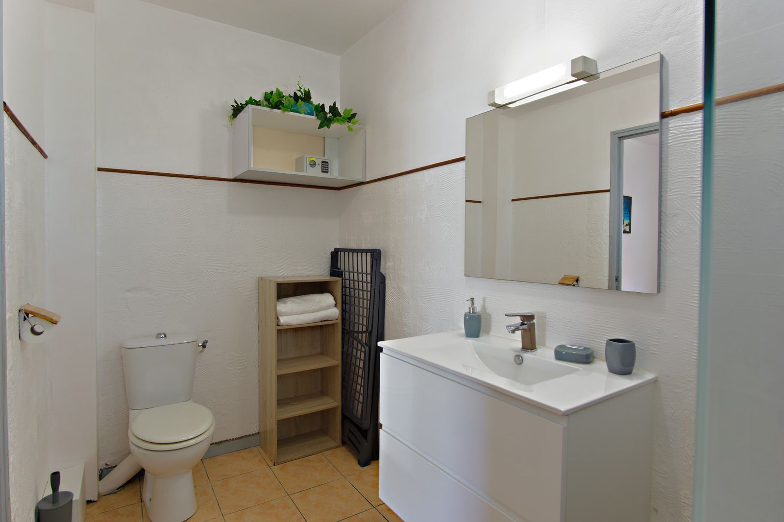A compact bathroom with a toilet, a sink, a mirror, a shelf with toiletries, and a small plant.
