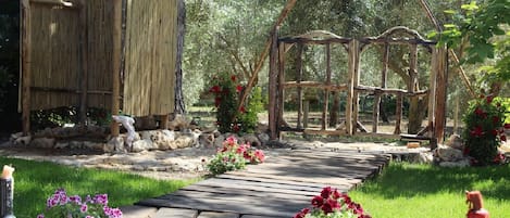 A wooden pathway leading to a rustic wooden structure with a thatched roof, surrounded by greenery and flowering plants.
