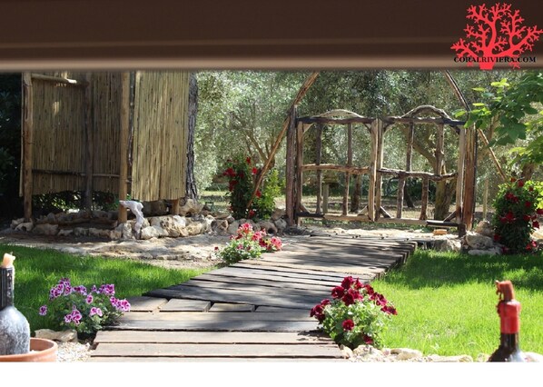 Un sendero de madera conduce a una estructura rústica de madera con techo de paja, rodeada de follaje y plantas florecientes.