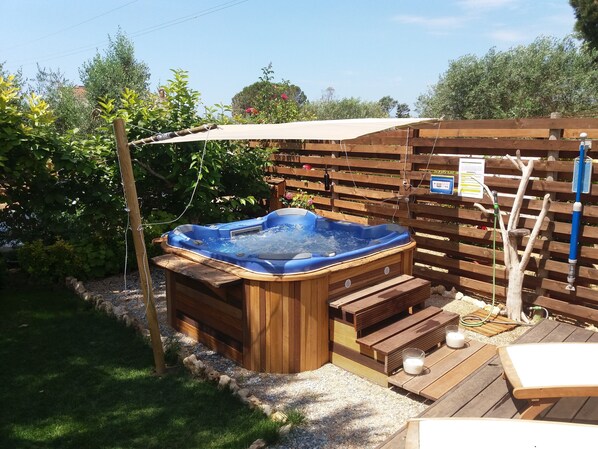 Un jacuzzi en un patio trasero con terraza de madera, rodeado de follaje y una valla de madera.