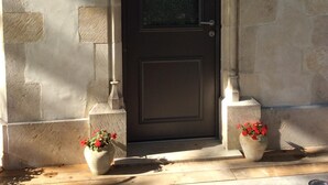 A stone building with a dark brown door, two potted plants with red flowers, and a small window above the door.